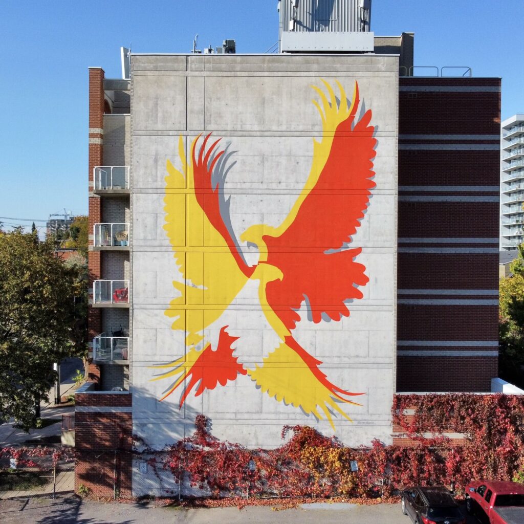 Silhouette image of two flying eagles painted on a large concrete wall. The eagles are red and yellow and where their wings overlap is left unpainted. There is a striped red brick building in the background.